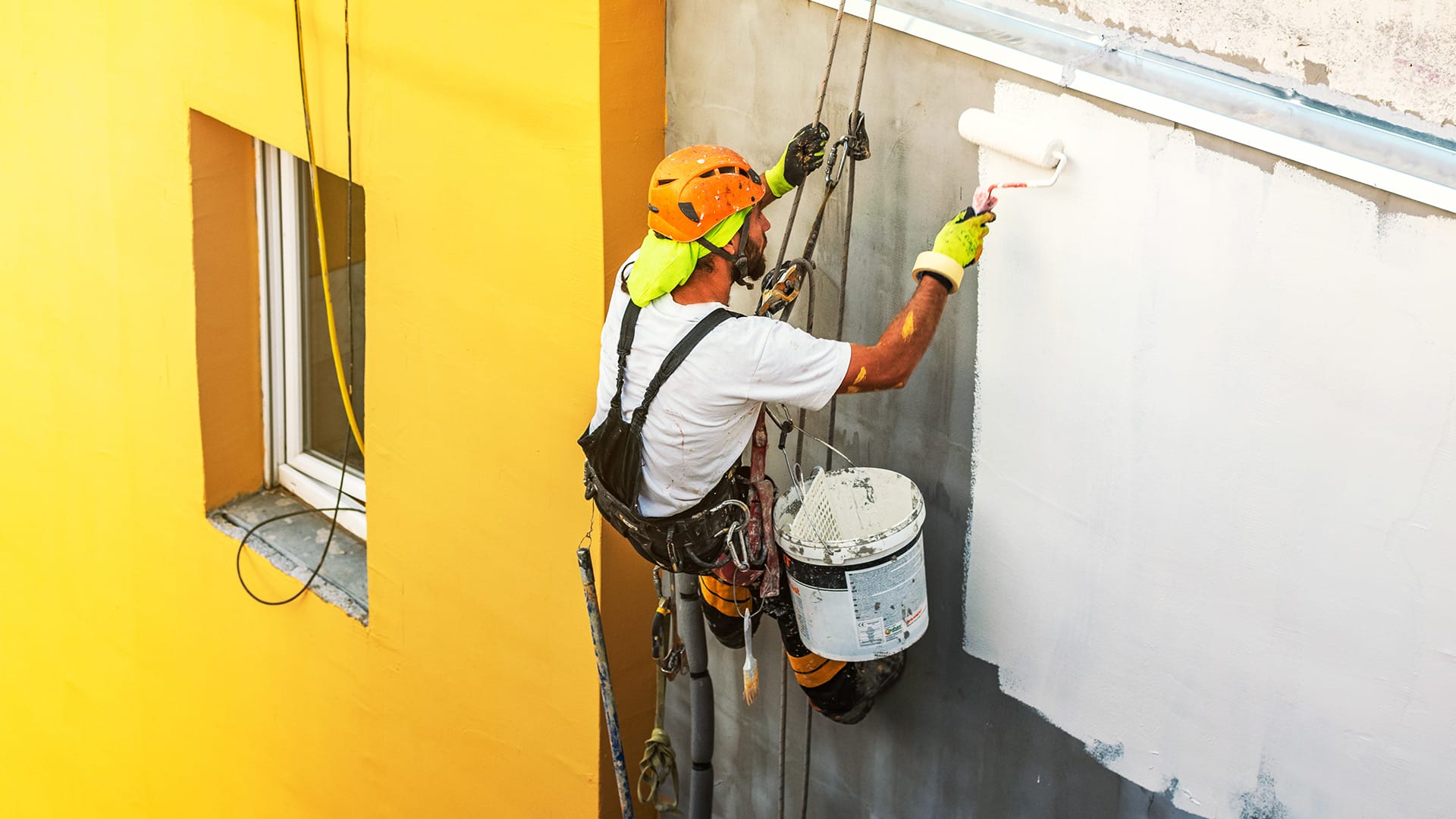 Industrial Painter on side of building with a white paint covered roller brush, next to a mustard yellow wall with a recessed window.