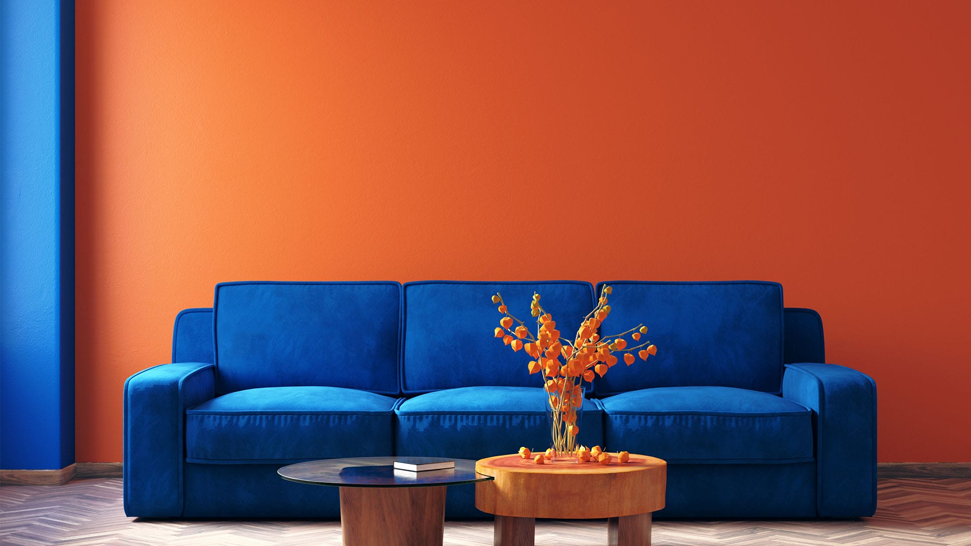 A modern living room featuring a vibrant blue sofa against an orange wall, with a wooden coffee table holding a vase of orange flowers in the center.