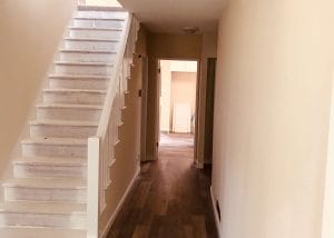 An empty hallway with a light colored staircase on the left and a doorway to another room at the end.